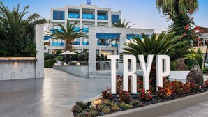 L’entrée d’un hôtel, avec une grande enseigne « TRYP », un bâtiment moderne aux fenêtres en verre bleu et un espace paysager agrémenté de plantes et d’un palmier.