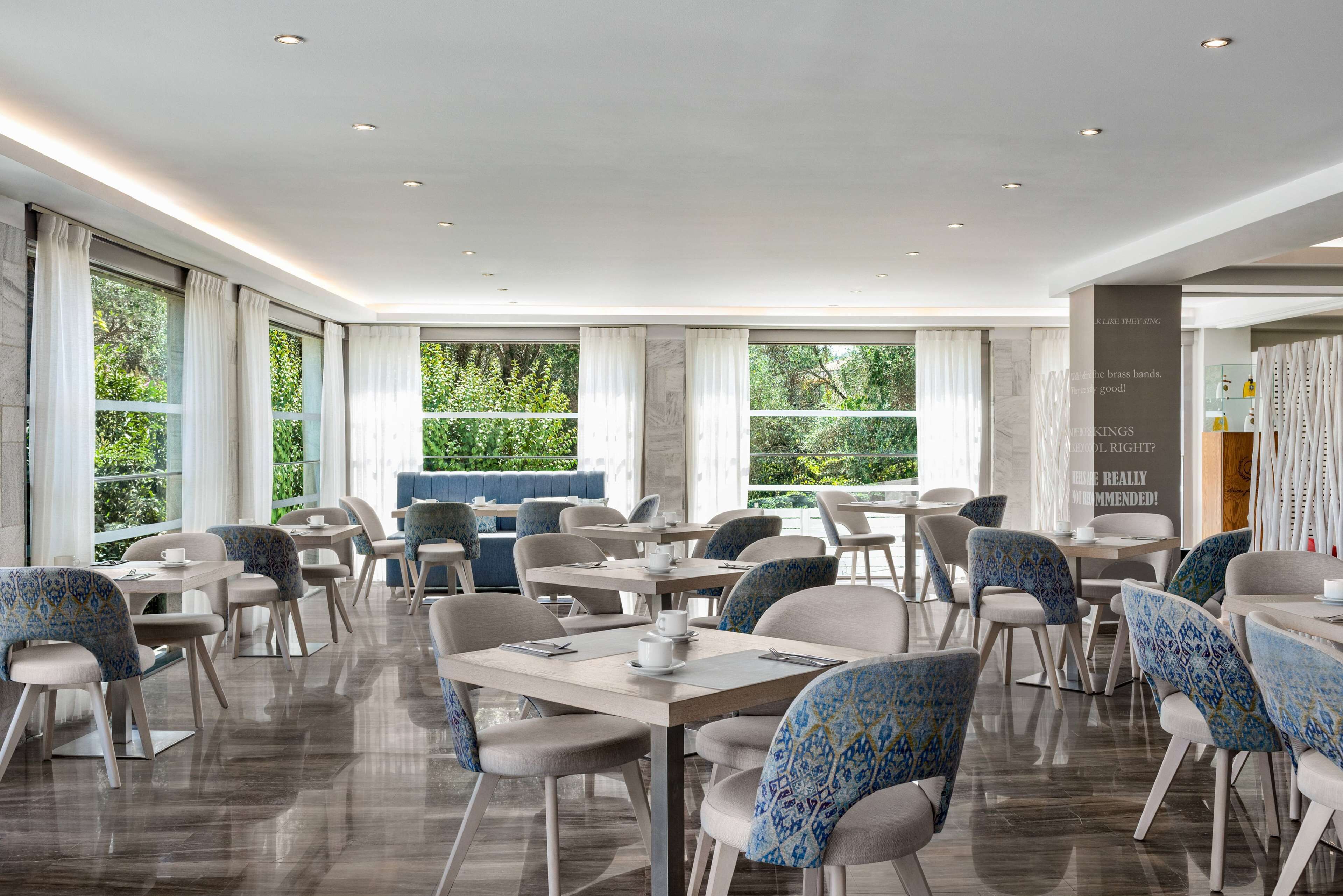 A modern dining area with tables and chairs, large windows, and a view of greenery outside.