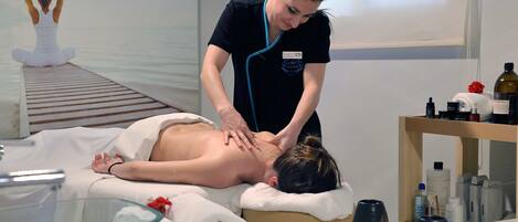 En massagebehandling i en spa-miljö, där klienten ligger på en behandlingsbänk, en terapeut utför behandlingen och olika bekvämligheter som erbjuds på spaanläggningen syns.