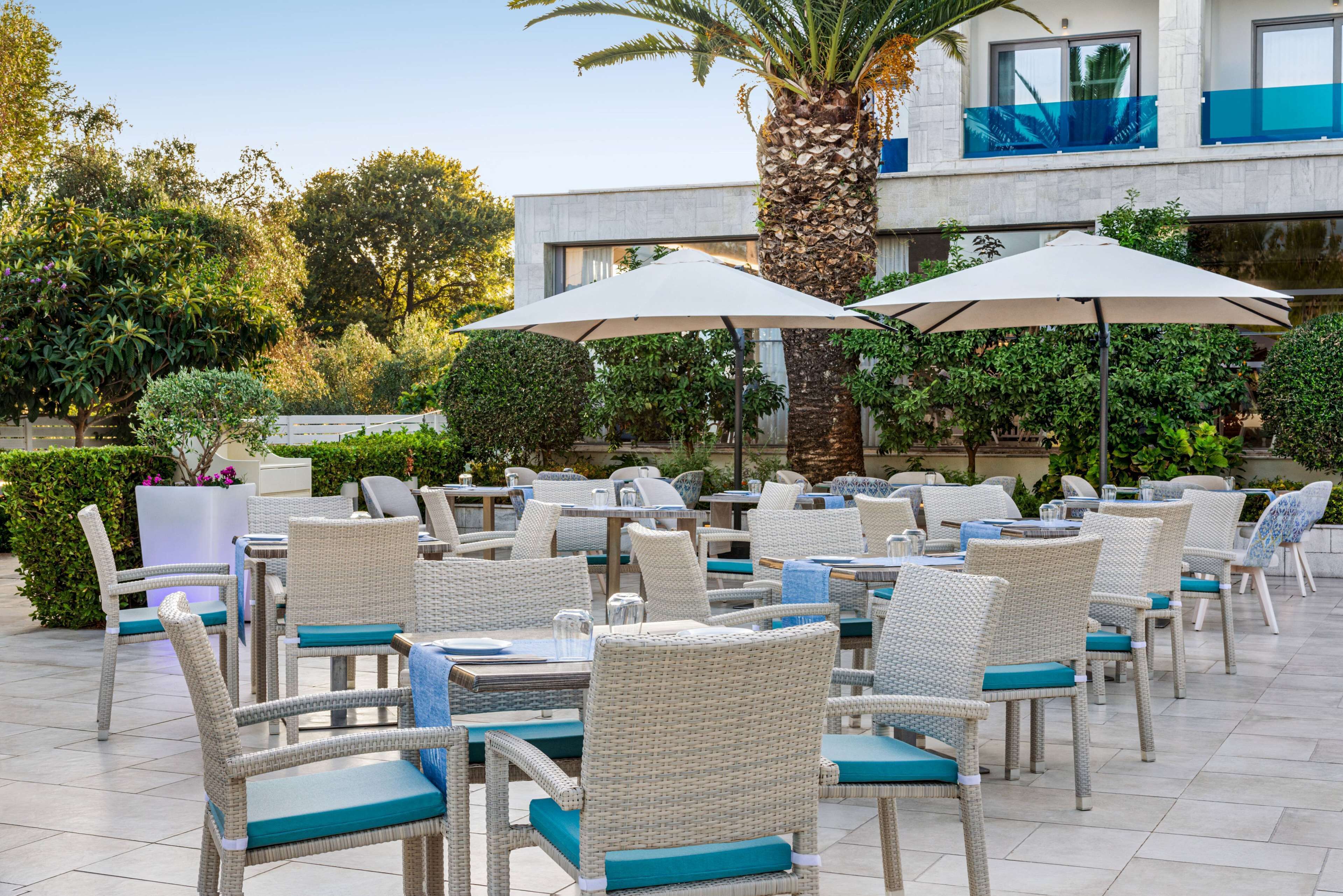 An outdoor seating area with wicker chairs and tables, surrounded by greenery and a modern building in the background.