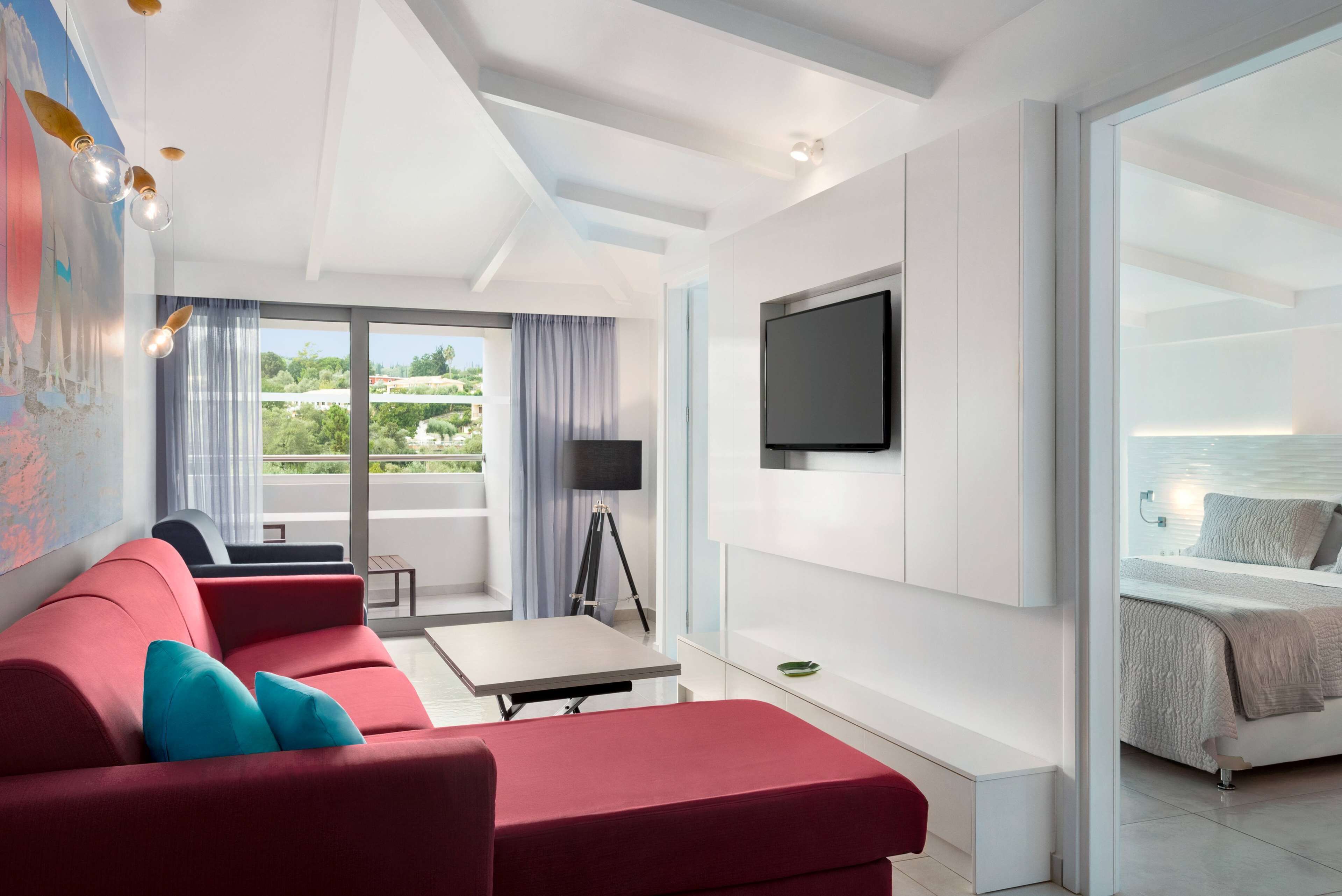 Una habitación de hotel moderna con un sofá rojo, un televisor de pantalla plana, un balcón con vistas a zonas verdes y un ventilador de techo.