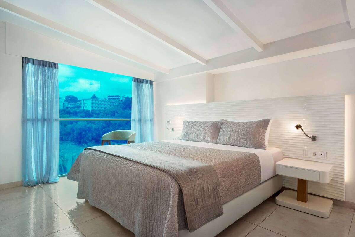 Een moderne slaapkamer met een groot bed, een nachtkastje en uitzicht op een stadsgezicht door een groot raam.