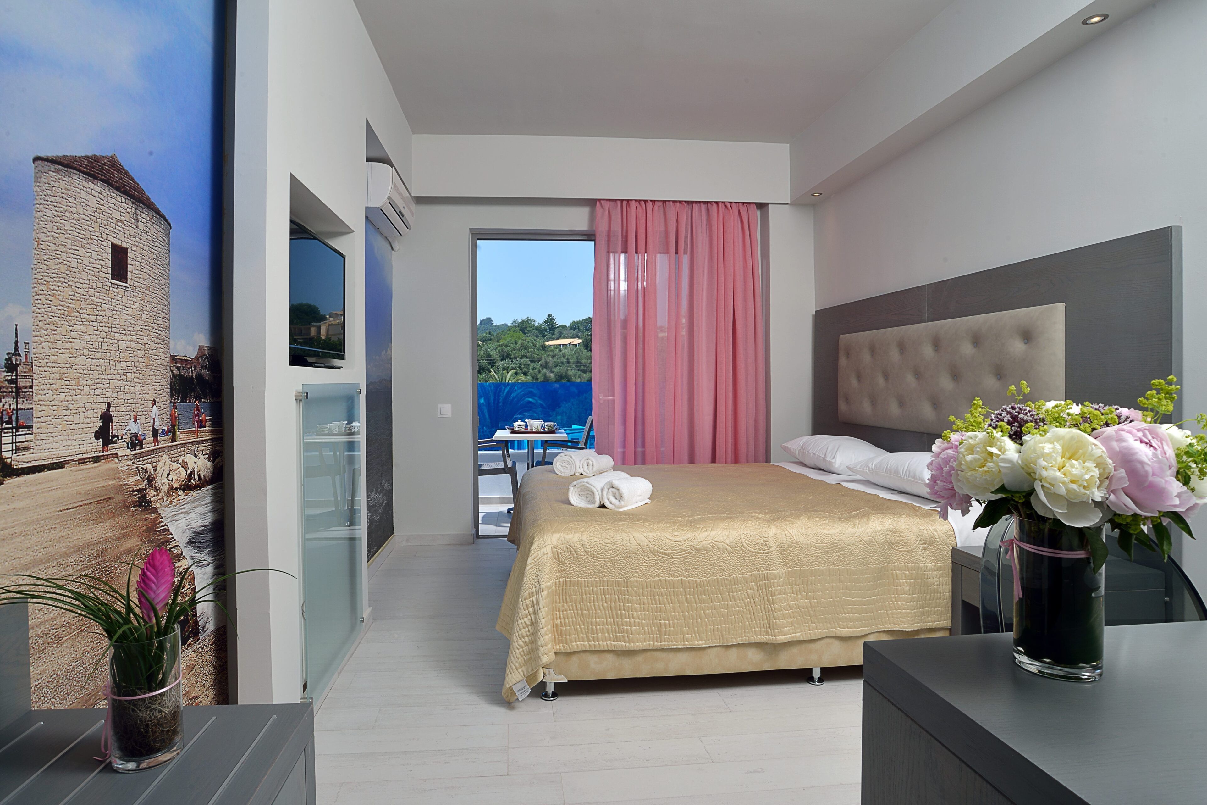 A modern hotel room with a large bed, a bedside table with a vase of flowers, a view of a coastal area, and a balcony with a table and chairs.