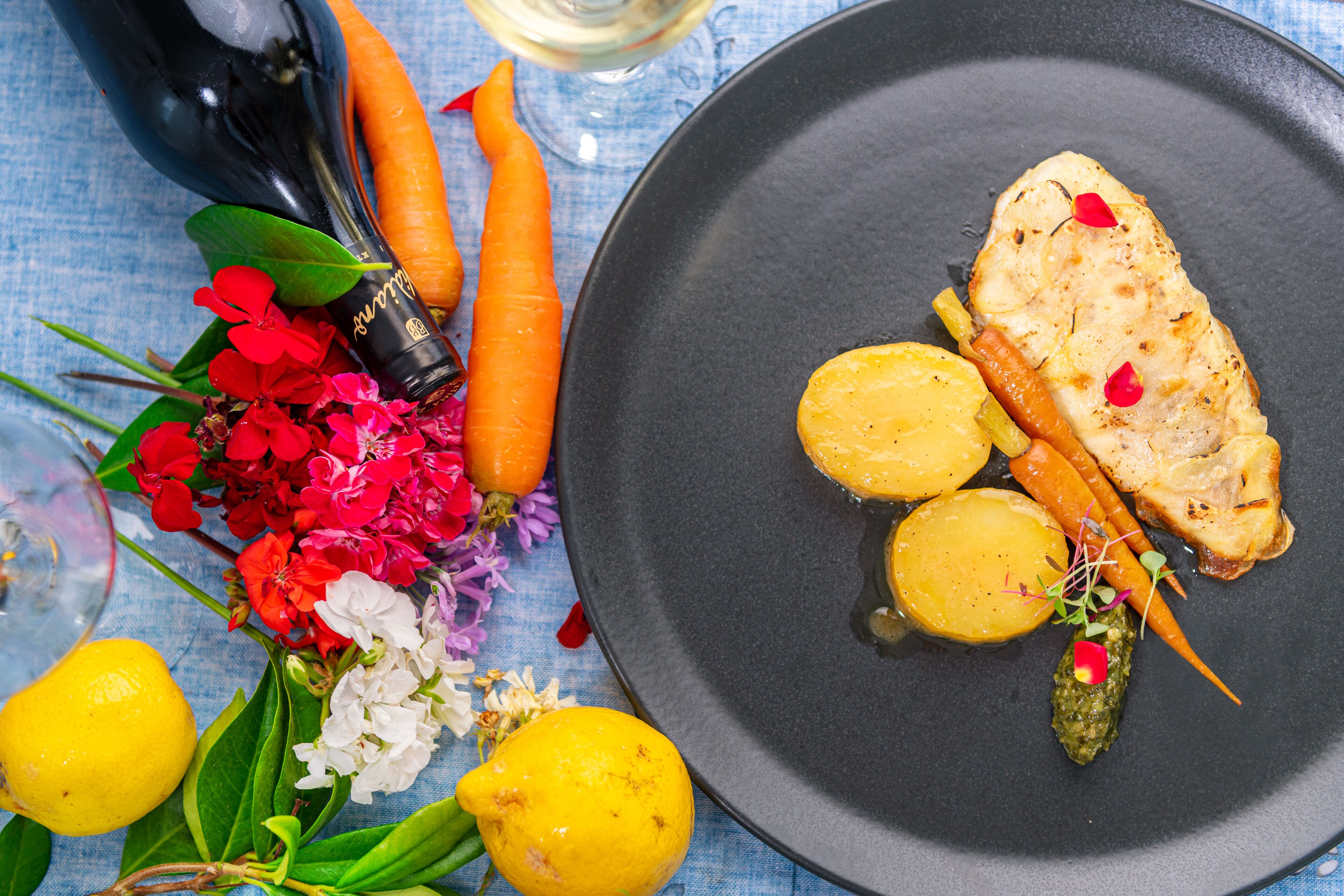 Un piatto con pesce, carote e patate, circondato da fiori e una bottiglia.