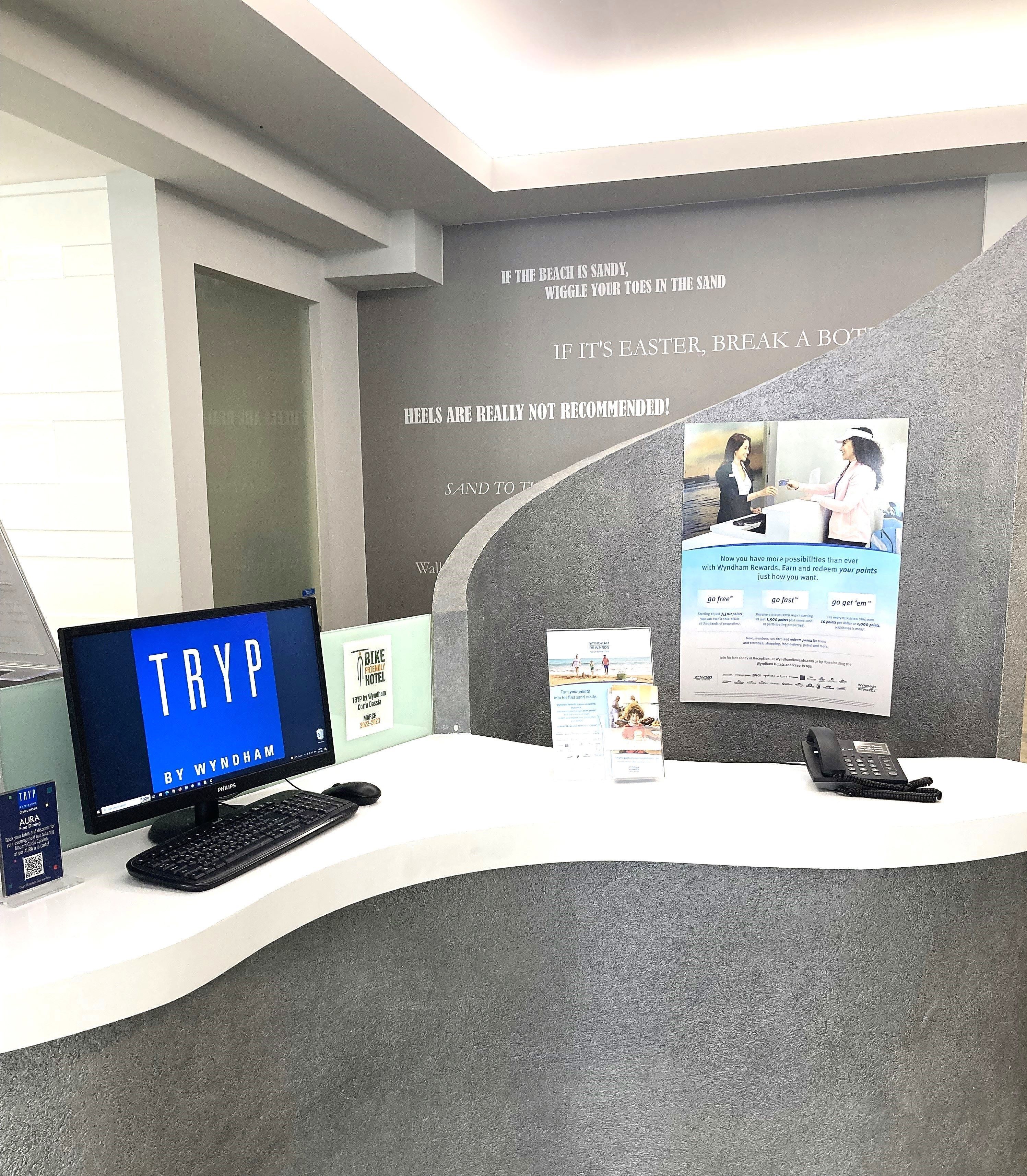 A reception desk with a computer monitor displaying 'TRYP BY WYNDHAM', a phone, and informational posters.