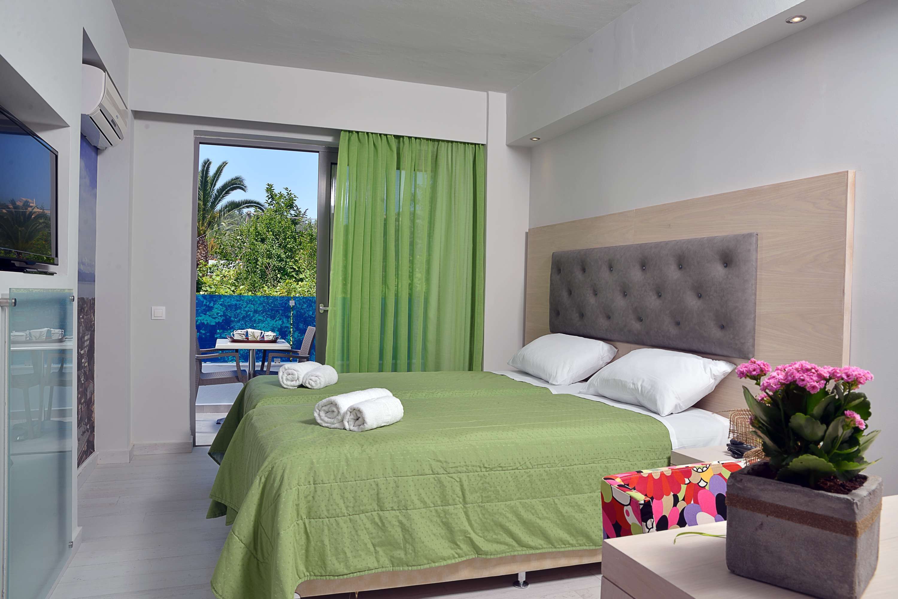 A modern bedroom with a green bedspread, a grey tufted headboard, white pillows, and a potted plant with pink flowers.