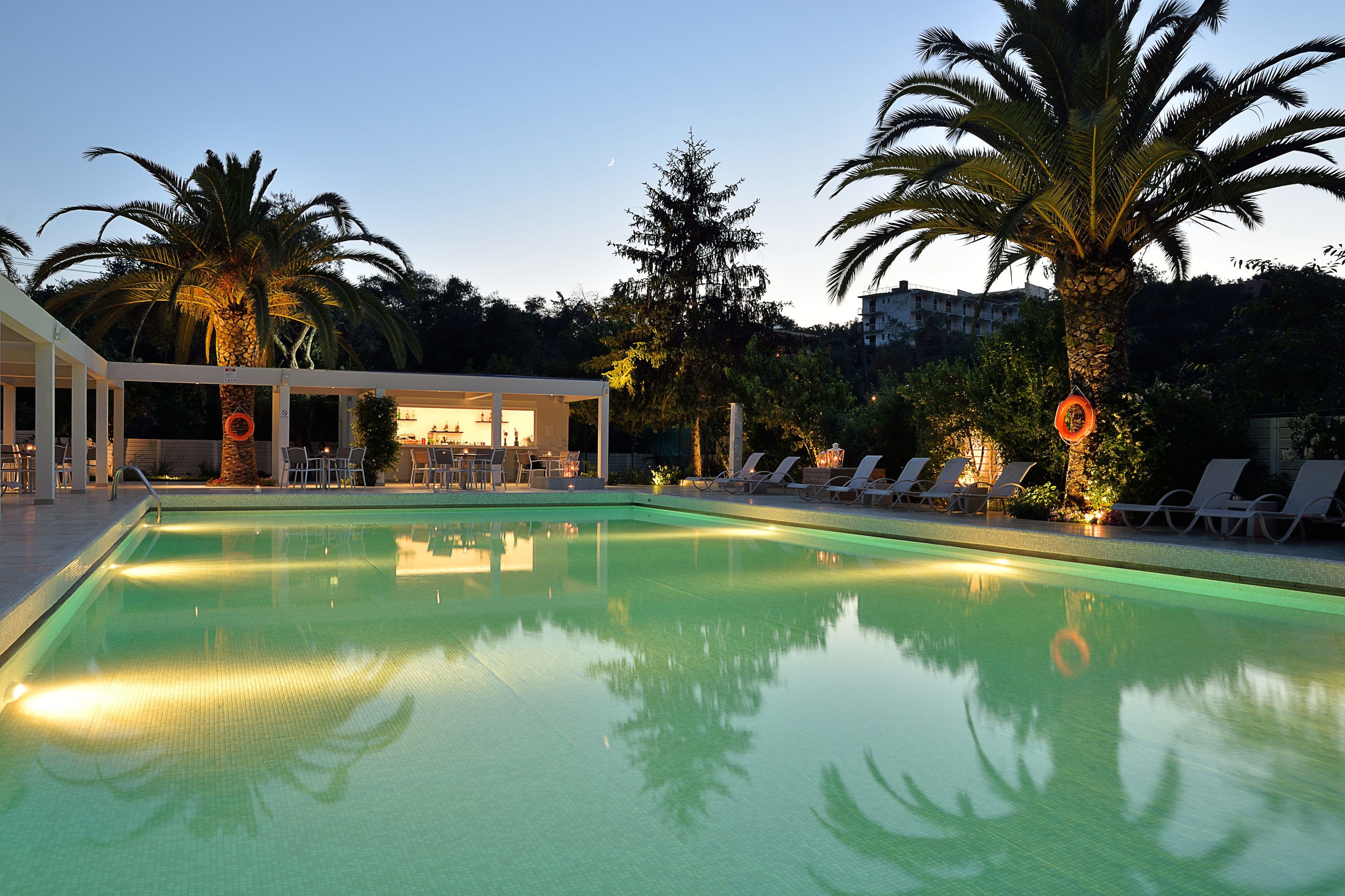 Piscina con sillones de descanso y palmeras, rodeada por un edificio con mesas al aire libre.