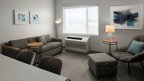 A modern living room with a gray sofa, two armchairs, a round coffee table, and a side table with a lamp. There is a large window providing natural light and a framed abstract painting on the wall. - Towneplace Suites Anchorage Midtown (Anchorage)