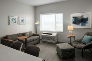 A modern living room with a gray sofa, two armchairs, a round coffee table, and a side table with a lamp. There is a large window providing natural light and a framed abstract painting on the wall. - Towneplace Suites Anchorage Midtown (Anchorage)