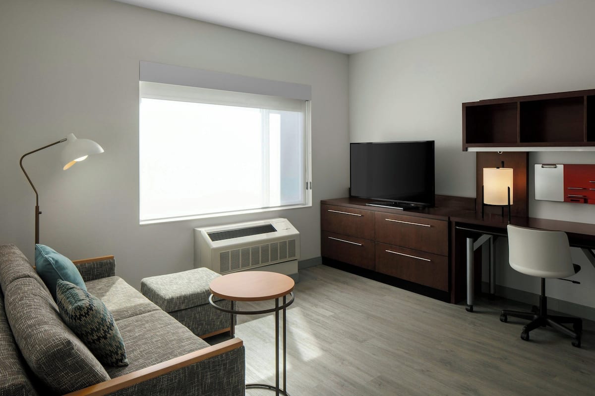 A compact living space with a sofa, a small round table, a desk with a chair, a television, and a window providing natural light.