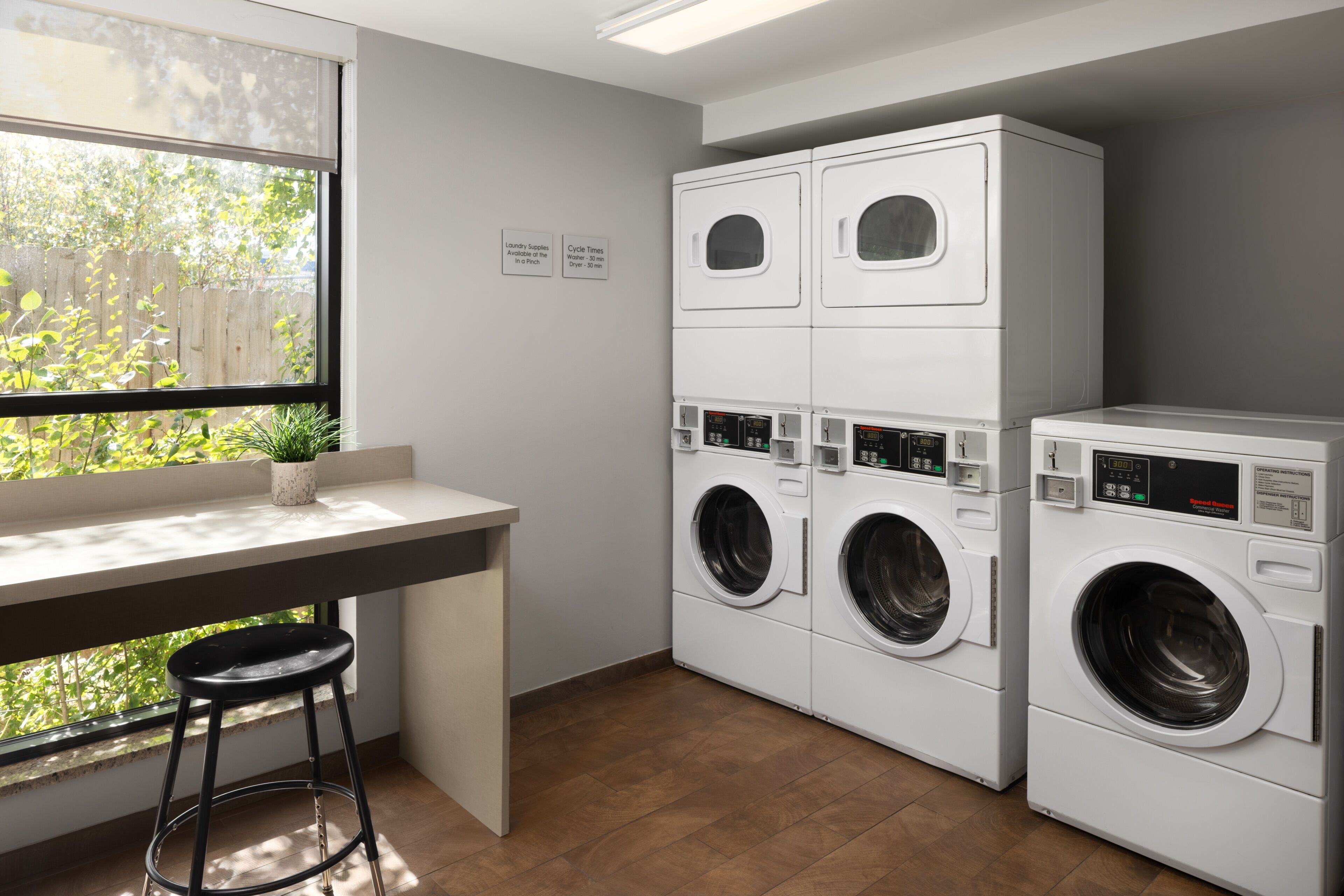 A laundry room with stacked washers and dryers, a window with a view of greenery, and a small bar stool.