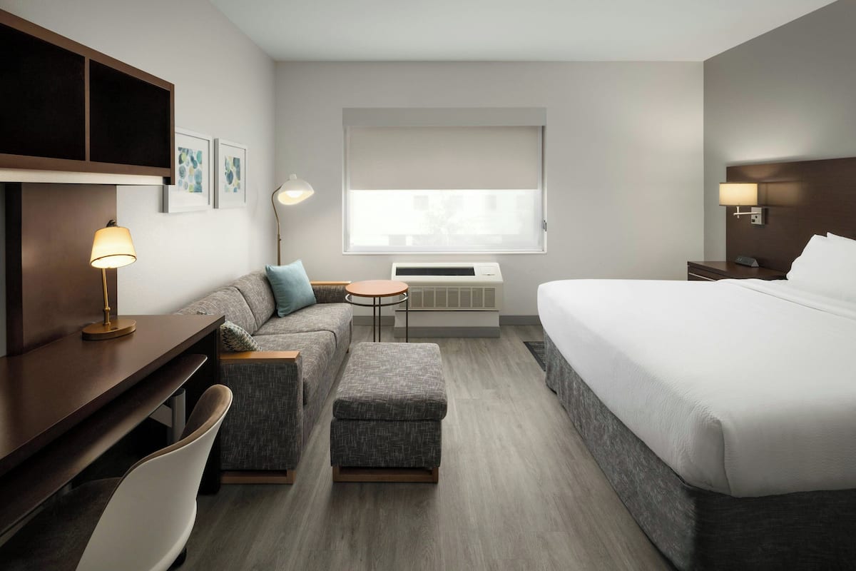 A modern hotel room with a bed, a sofa, a desk, a chair, a small table, and a lamp.