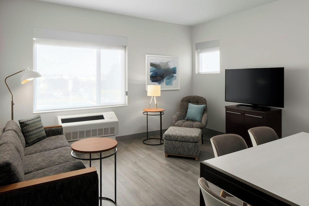 A modern hotel room with a sofa, armchair, round table, and a flat-screen TV.