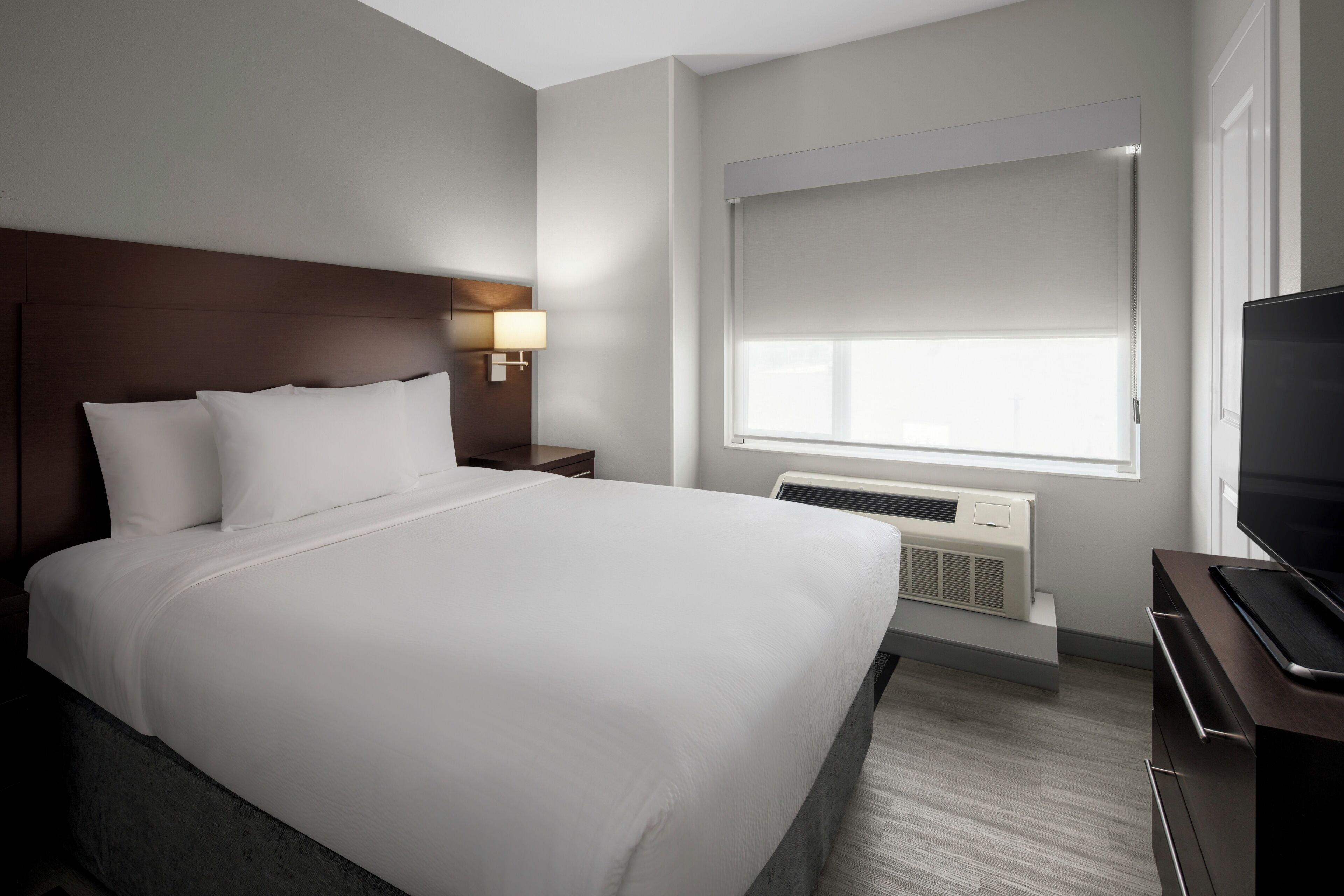 Una habitación de hotel con una cama grande, una mesita de noche con lámpara, una ventana con persianas, un radiador y un televisor sobre un soporte.