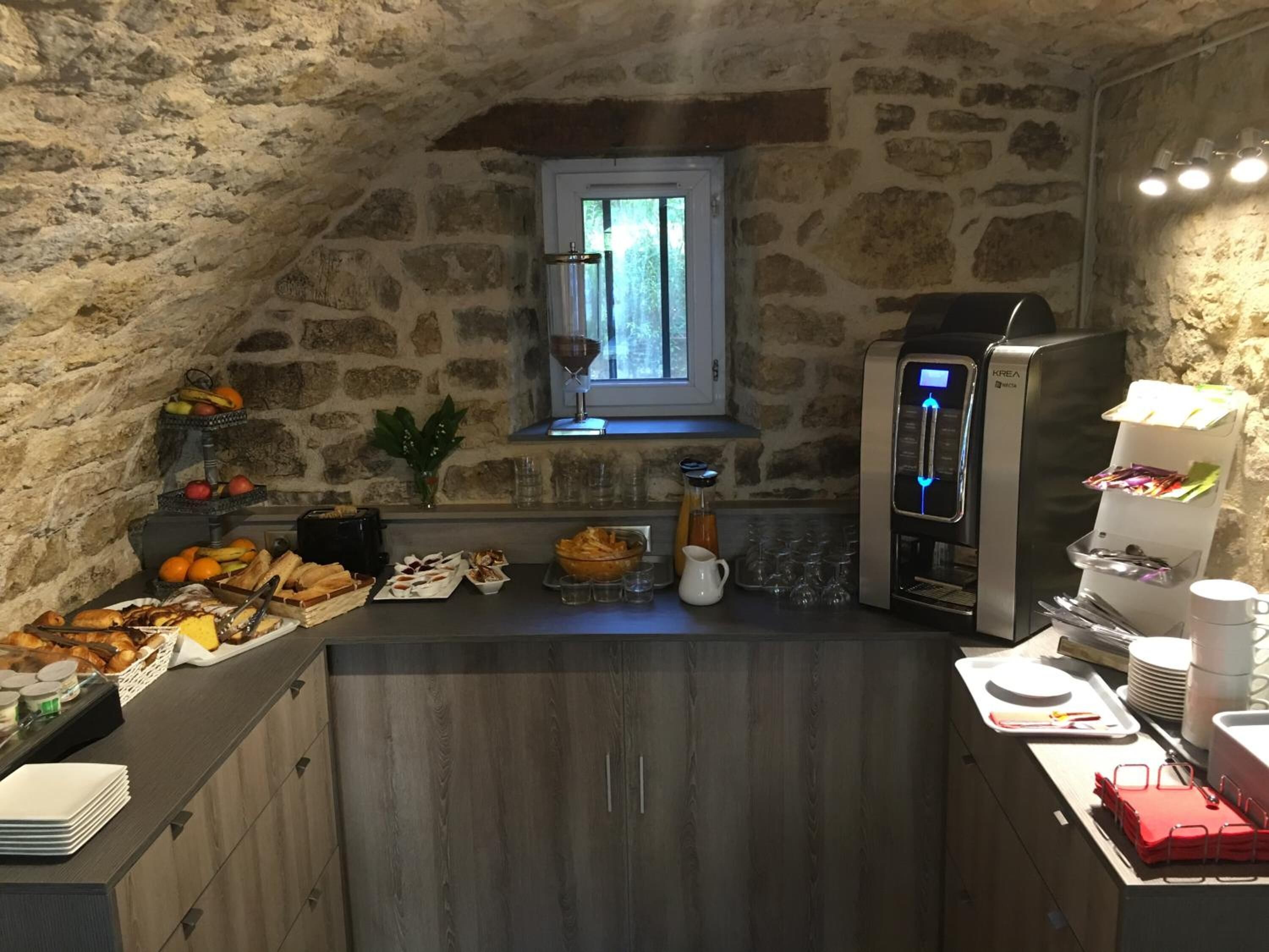 Une cuisine avec des murs en pierre, une fenêtre, une machine à café et divers aliments disposés sur le plan de travail.