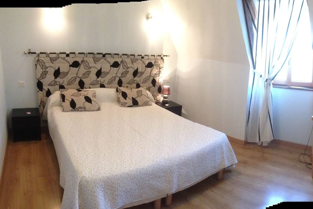 A bedroom with a bed, a nightstand, a lamp, a chair, and a window with curtains.