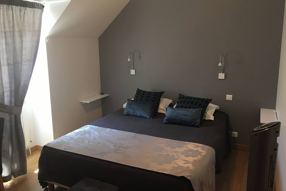 A bedroom with a bed, two bedside tables, a nightstand, a bench, a window with curtains, and a sloped ceiling.