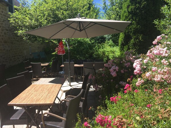 An outdoor patio with wooden tables and wicker chairs, a large umbrella, and blooming pink flowers. - Hôtel & Spa Rodier (Campagnac)