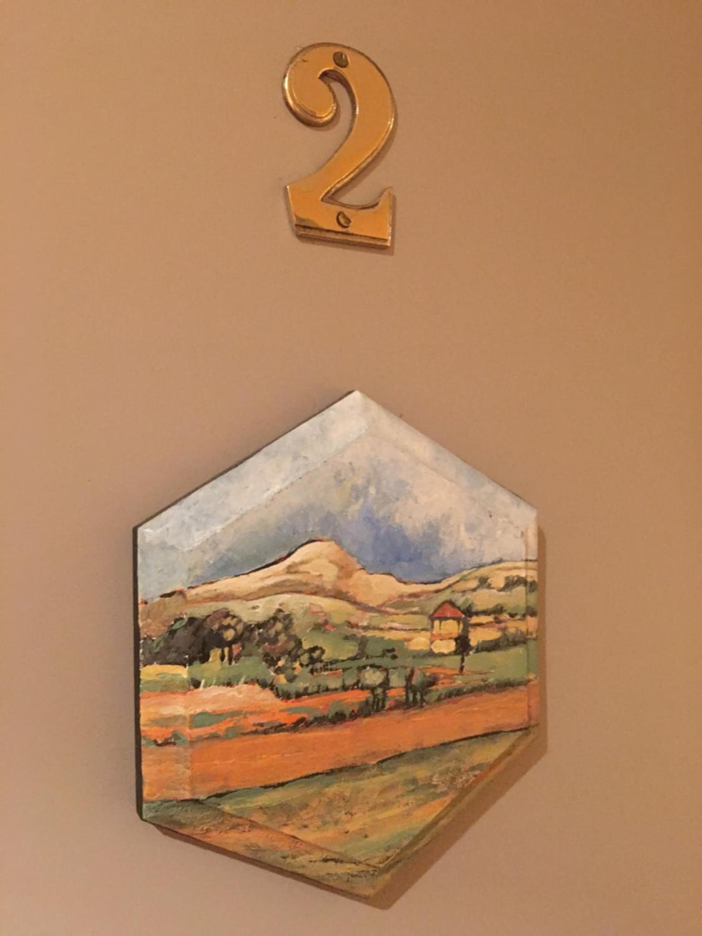A hexagonal painting of a rural landscape with a mountain and houses, mounted on a wall above a number '2'.