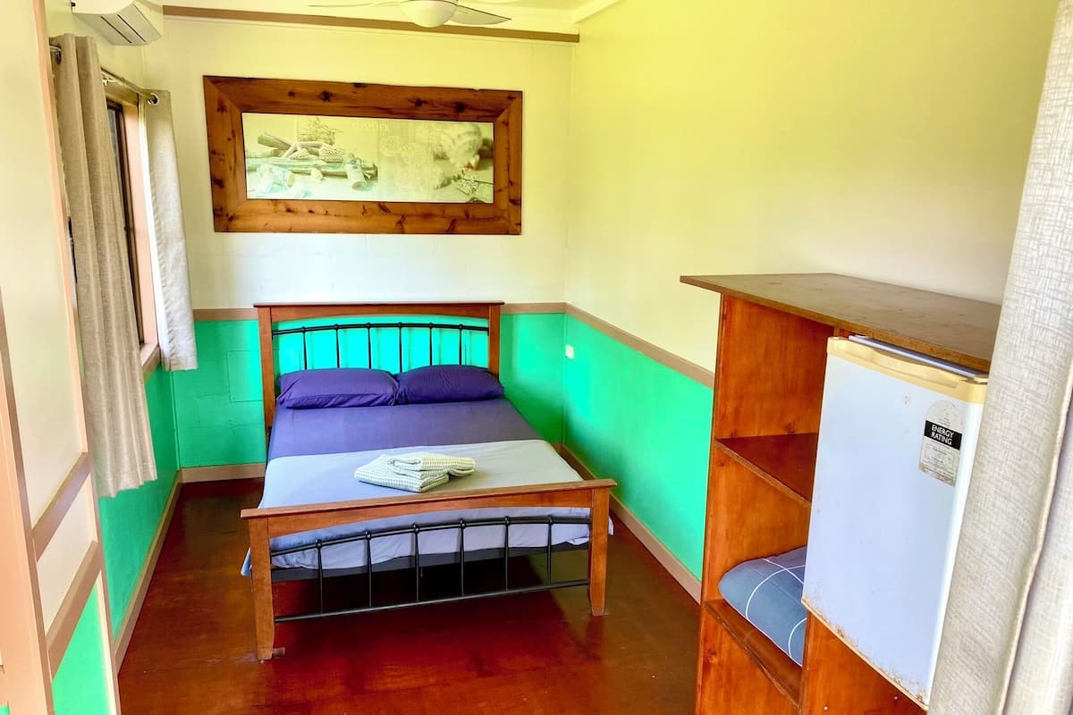 A small, compact room with a single bed, a wooden headboard, a small table, and a wall-mounted shelf.