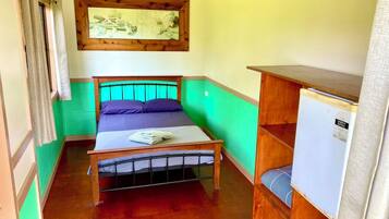Quarto pequeno e compacto com uma cama de solteiro, cabeceira de madeira, uma mesa pequena e uma prateleira fixada na parede.