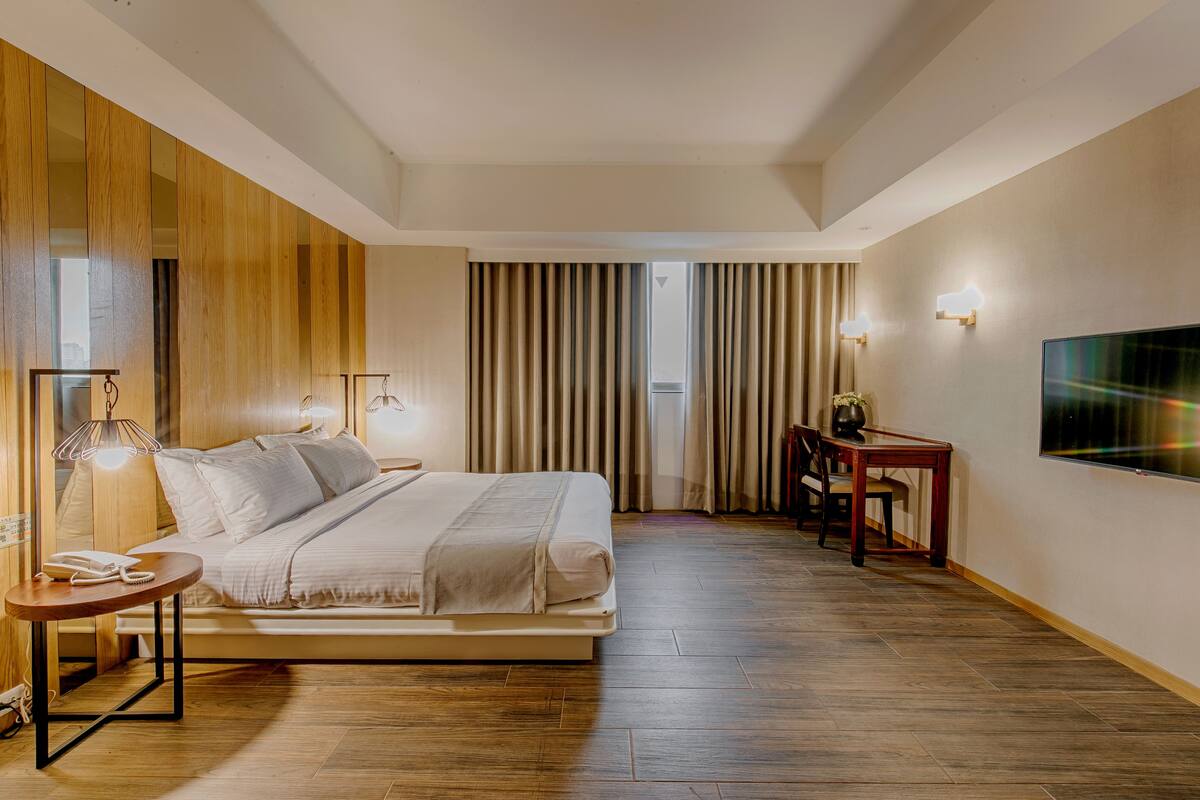 A modern hotel room with a bed, bedside tables, a desk, a chair, a TV, and curtains.