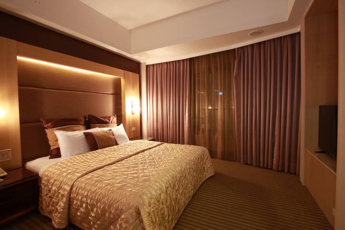 A hotel room with a large bed, a headboard with integrated lighting, a flat-screen TV, and a large window with curtains.