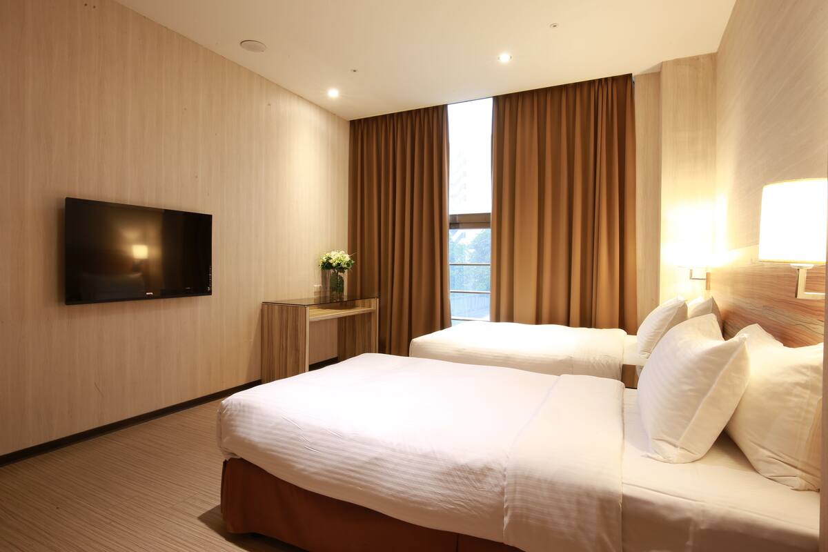 A hotel room with two beds, a flat-screen TV, a desk with a lamp, and a window with curtains.
