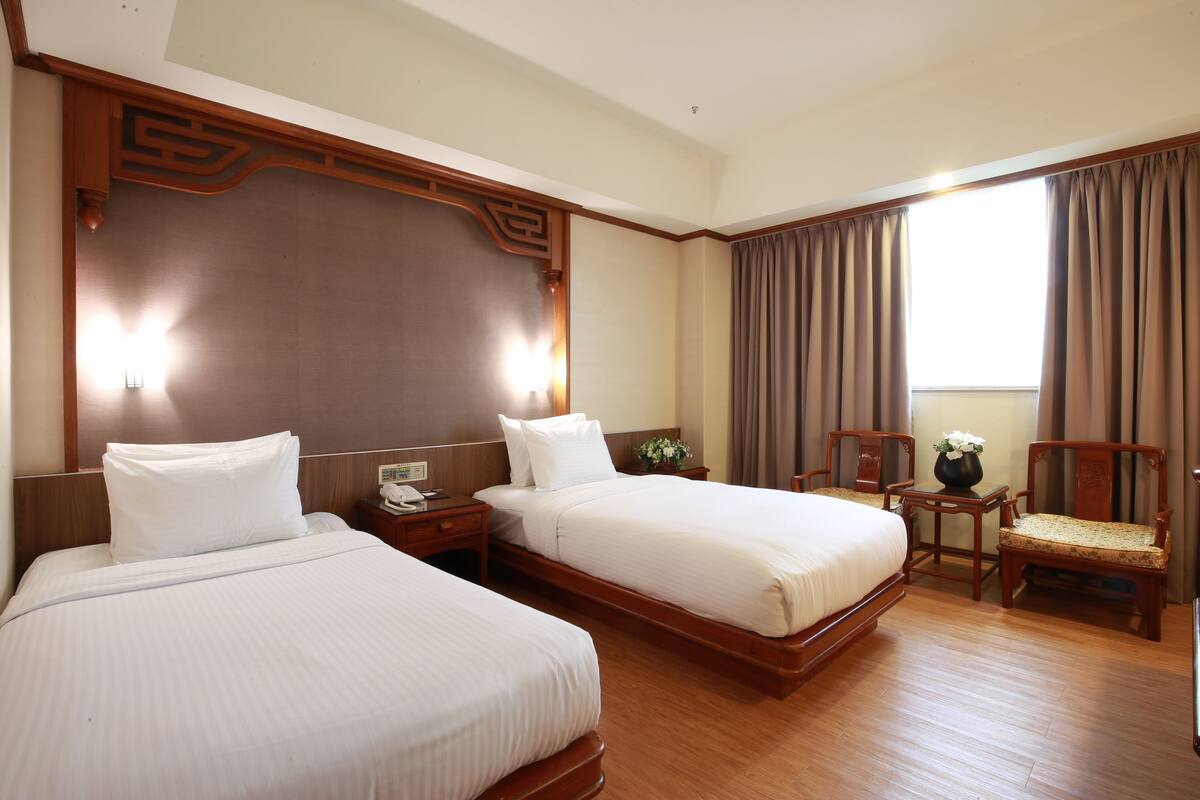 A hotel room with two beds, a wooden headboard, a nightstand, a chair, and a window with curtains.