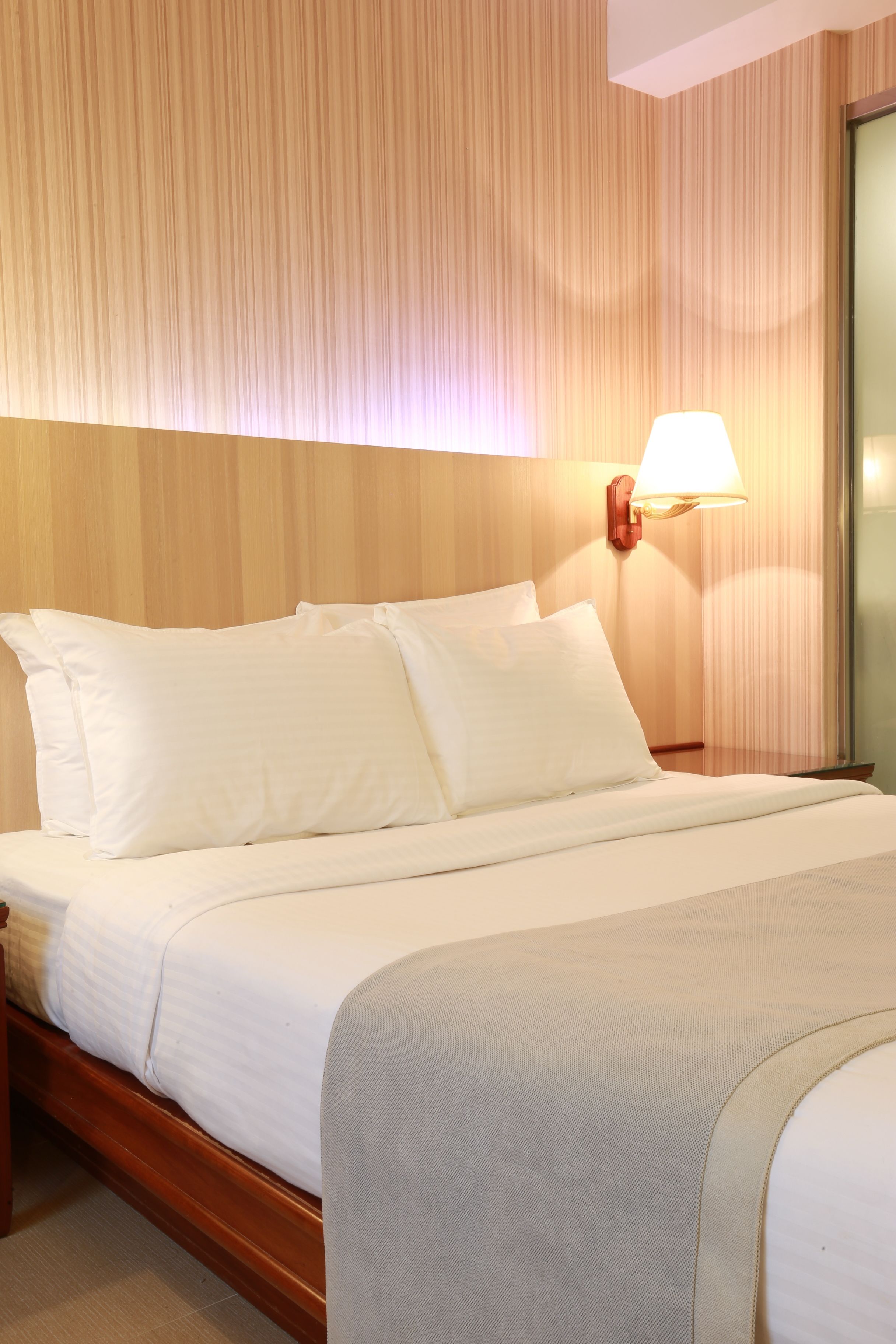 Una cama bien tendida con sábanas y almohadas blancas, un cabecero de madera y una lámpara fijada a la pared.