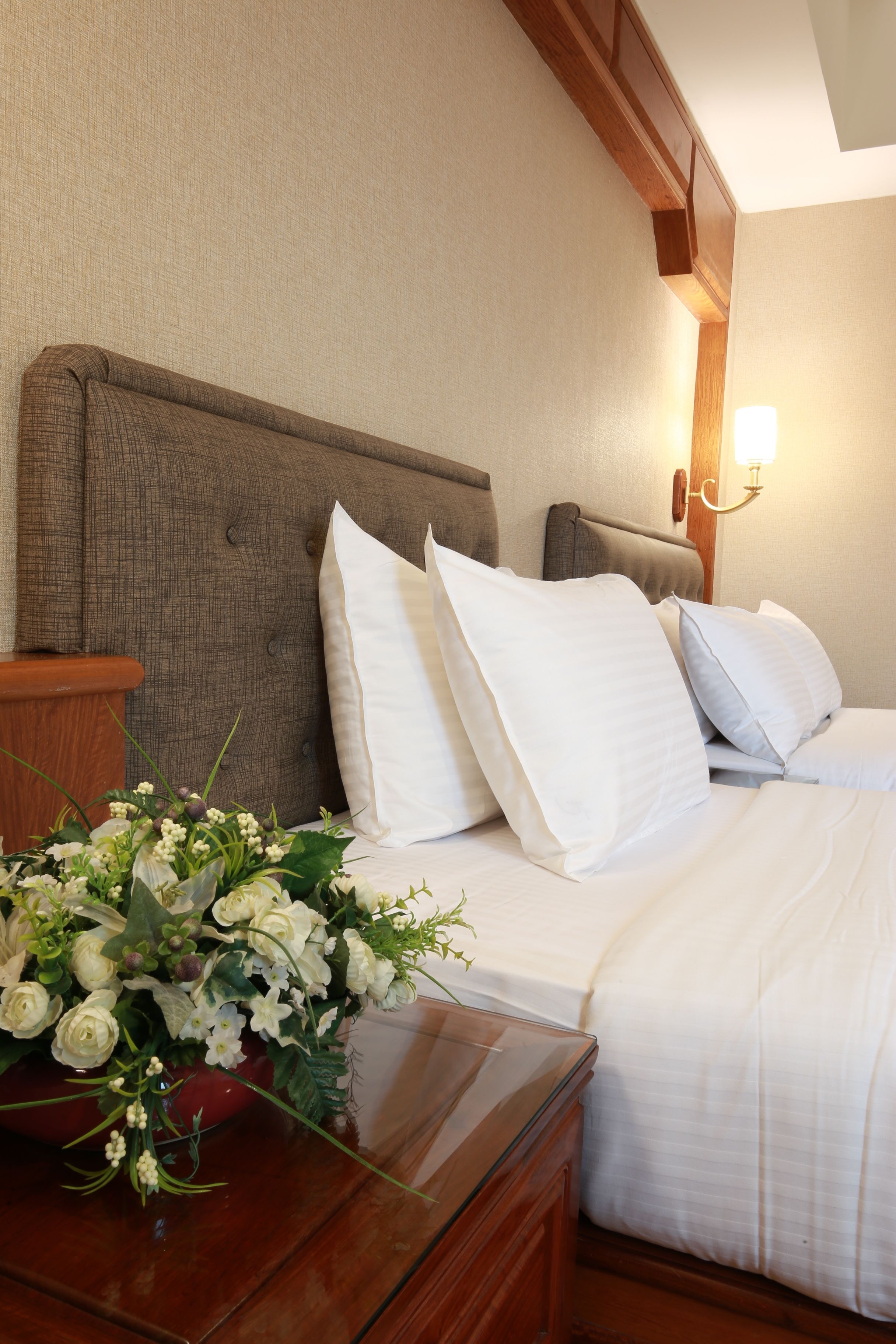 A hotel room with a bed, pillows, a nightstand, a vase of flowers, and a wall-mounted light fixture.