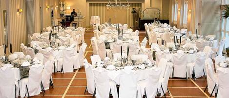 A banquet hall with round tables set for a formal event, featuring white tablecloths and chairs, and a chandelier in the center.