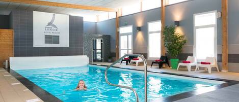 Une piscine intérieure avec un sol carrelé, un escalier métallique et une personne qui nage. On aperçoit des chaises longues et une plante en pot près de la piscine.