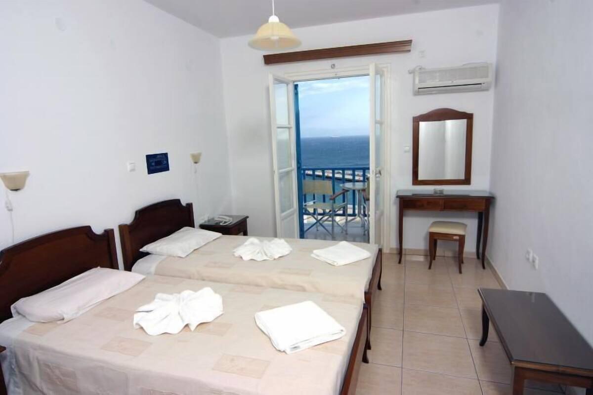 A hotel room with two beds, a desk, a chair, a mirror, and a view of the sea through an open door.