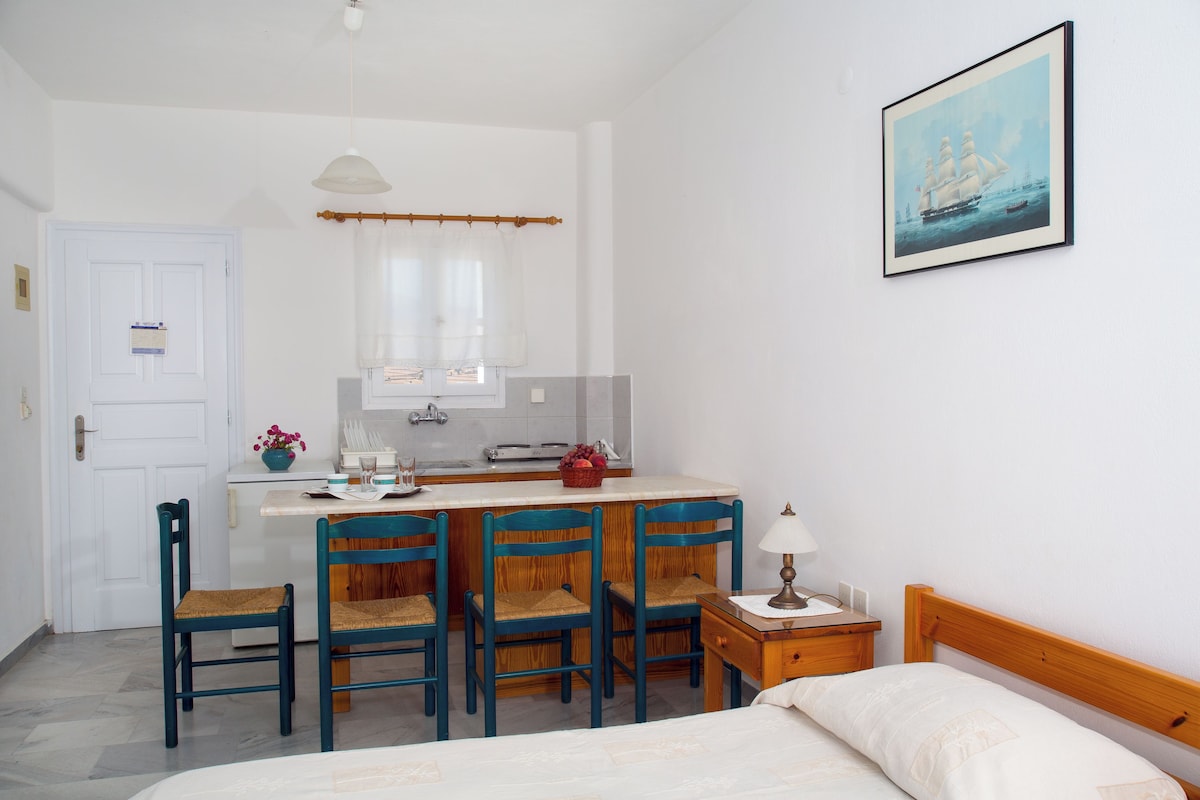 Una habitación con cama, un escritorio con sillas, una mesita de noche con lámpara, un cuadro en la pared y una ventana con cortinas.