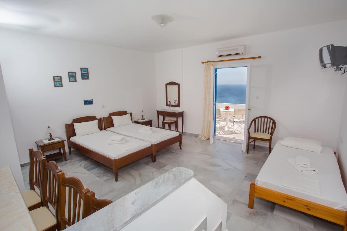 A bedroom with two beds, a wooden chair, a desk, and a view of the sea through an open door.
