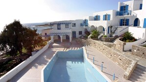 A pool area with a view of the sea, a white building with blue accents, and a stone wall. - Arkas Inn (Paros)