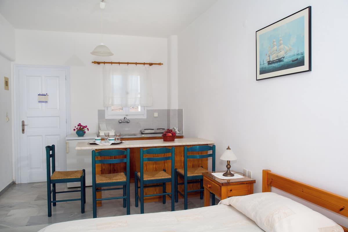 A room with a bed, a desk with chairs, a small table with a lamp, a painting on the wall, and a window with curtains.