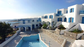 A pool area with a view of the sea, white buildings with blue accents, and stone steps leading to a covered area. - Arkas Inn (Paros)