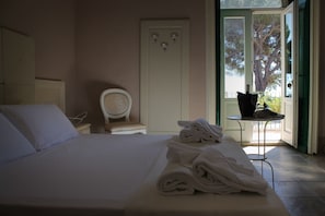 A hotel room with a bed, a chair, a table with towels, and a door leading to an outdoor area with a view of a tree. - Torre del Mare (Ispica)