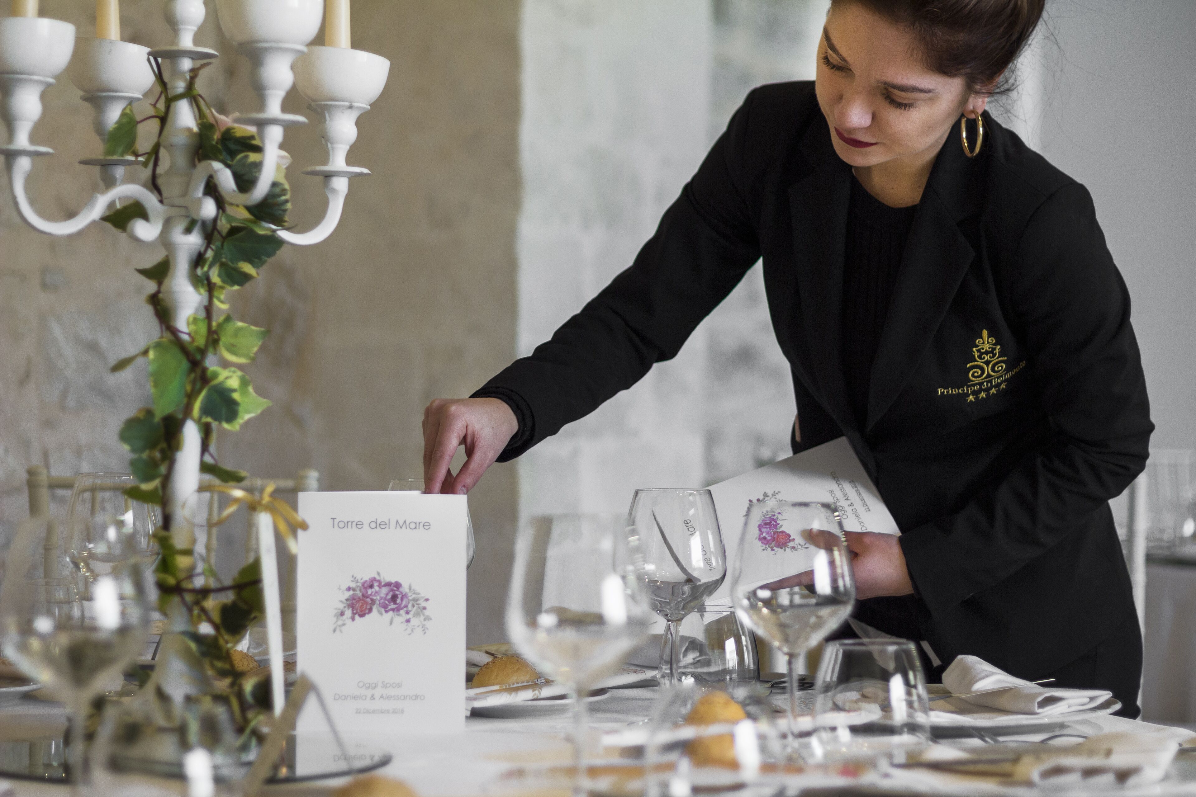 A person arranging a table with wine glasses and a menu.