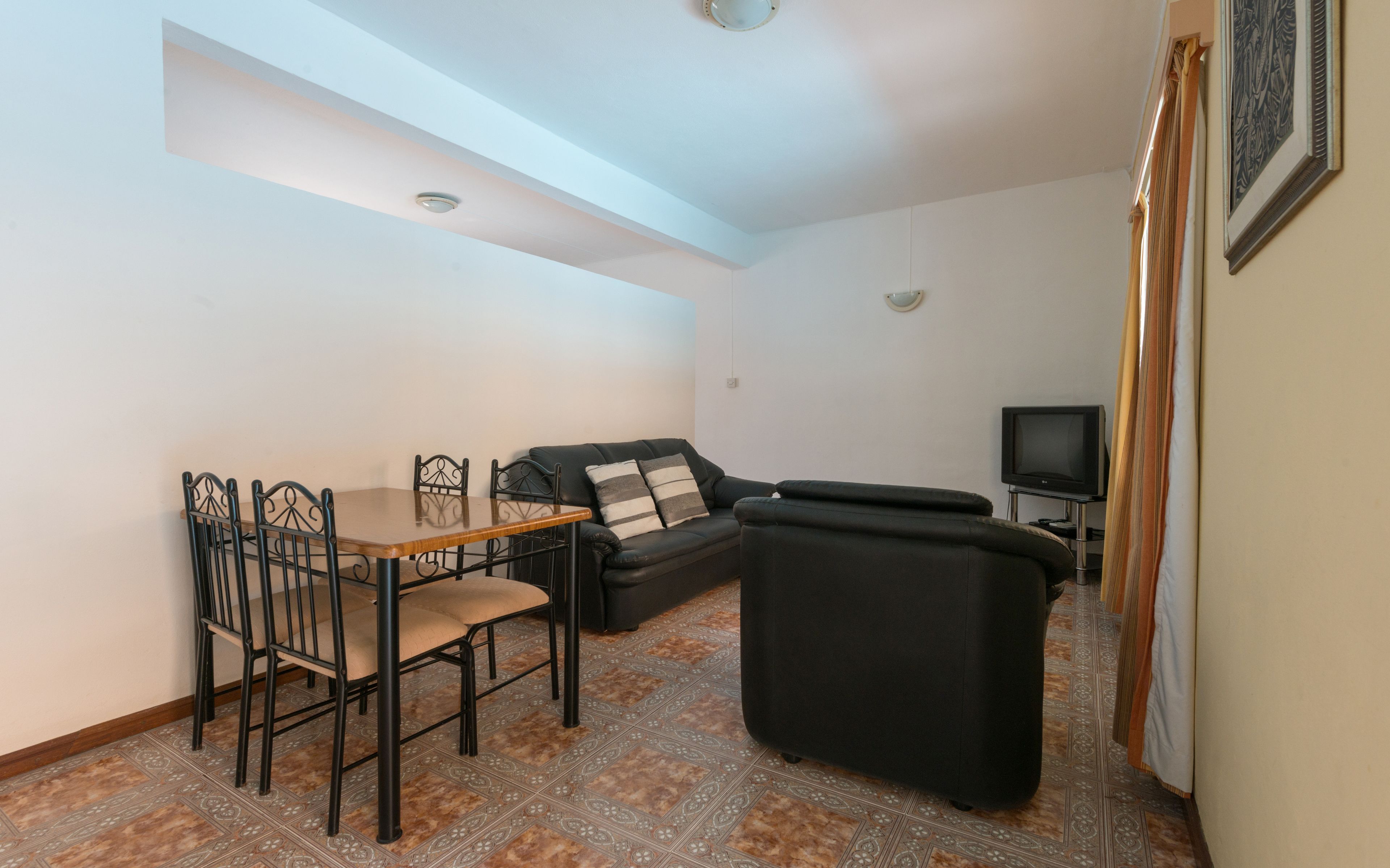 A living room with a sofa, a dining table with chairs, a television, and patterned floor tiles.