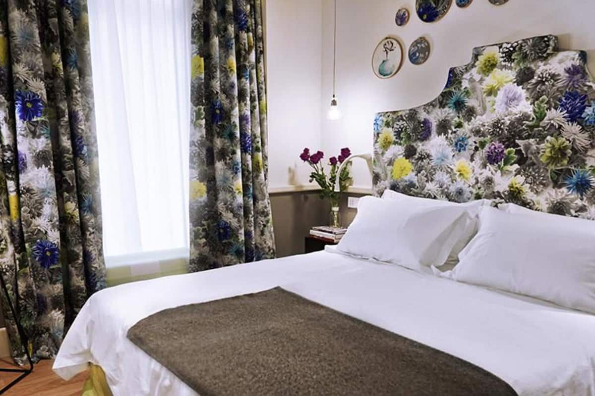 A hotel room with a bed, floral curtains, a bedside table with a vase of flowers, and a wall with decorative plates.