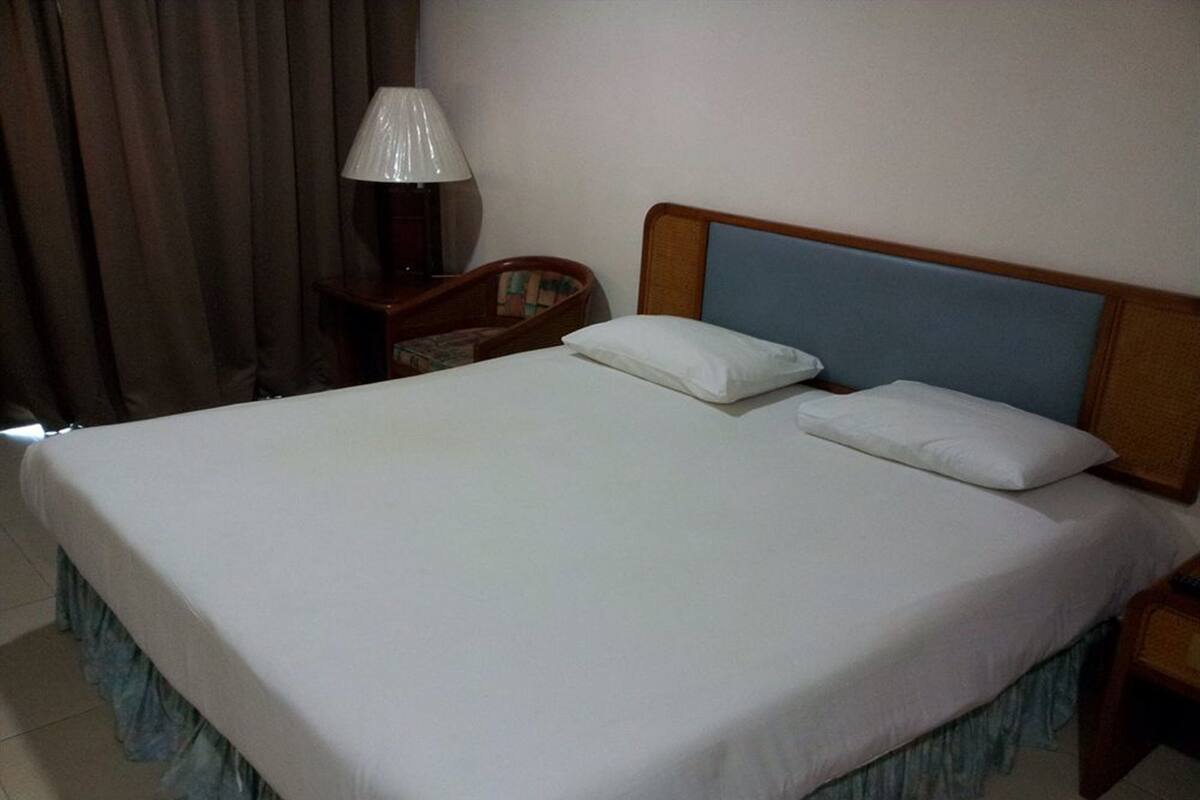 A neatly made bed with white linens and two pillows, a wooden headboard, a nightstand with a lamp, and a chair with a cushion.