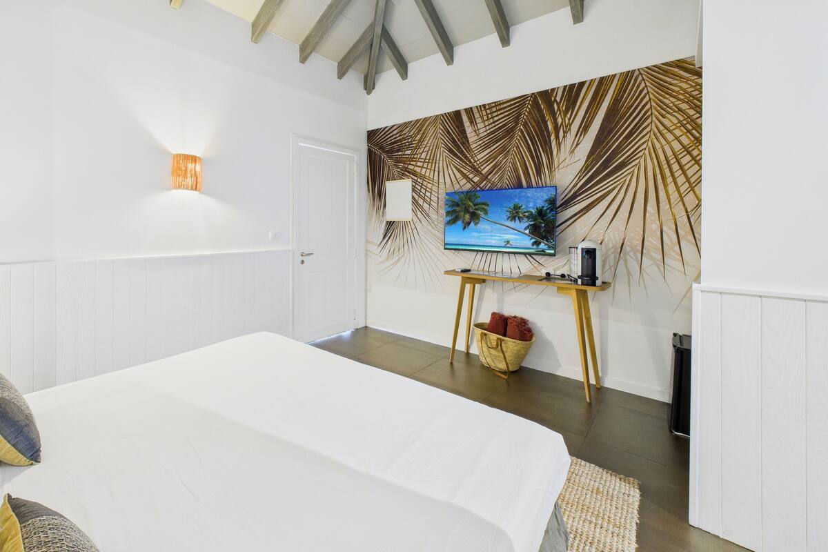 A bedroom with a bed, a TV on a stand, a lamp, and a wall with a palm leaf pattern.
