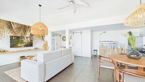 A modern living room with a sofa, dining table, and chairs. There is a TV mounted on the wall, a ceiling fan, and a large window. - Amnios St Barth (St. Barthelemy)