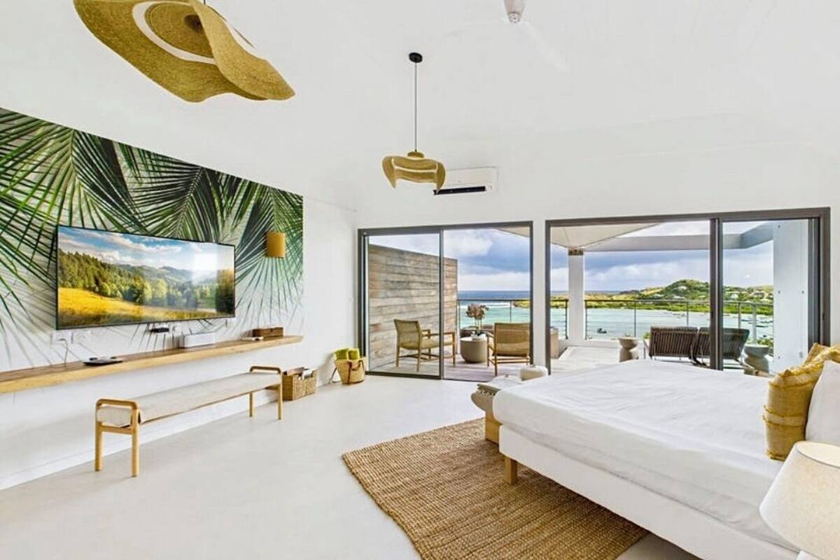 A modern bedroom with a large bed, a TV, and a balcony with a view of the sea.