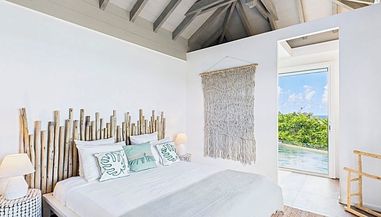 A bedroom with a wooden headboard, a large bed with multiple pillows, a nightstand with a lamp, and a view of a swimming pool through an open door.