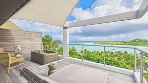 A balcony with a wooden floor, a sofa, a chair, a hot tub, and a view of a turquoise sea and green landscape. - Amnios St Barth (St. Barthelemy)