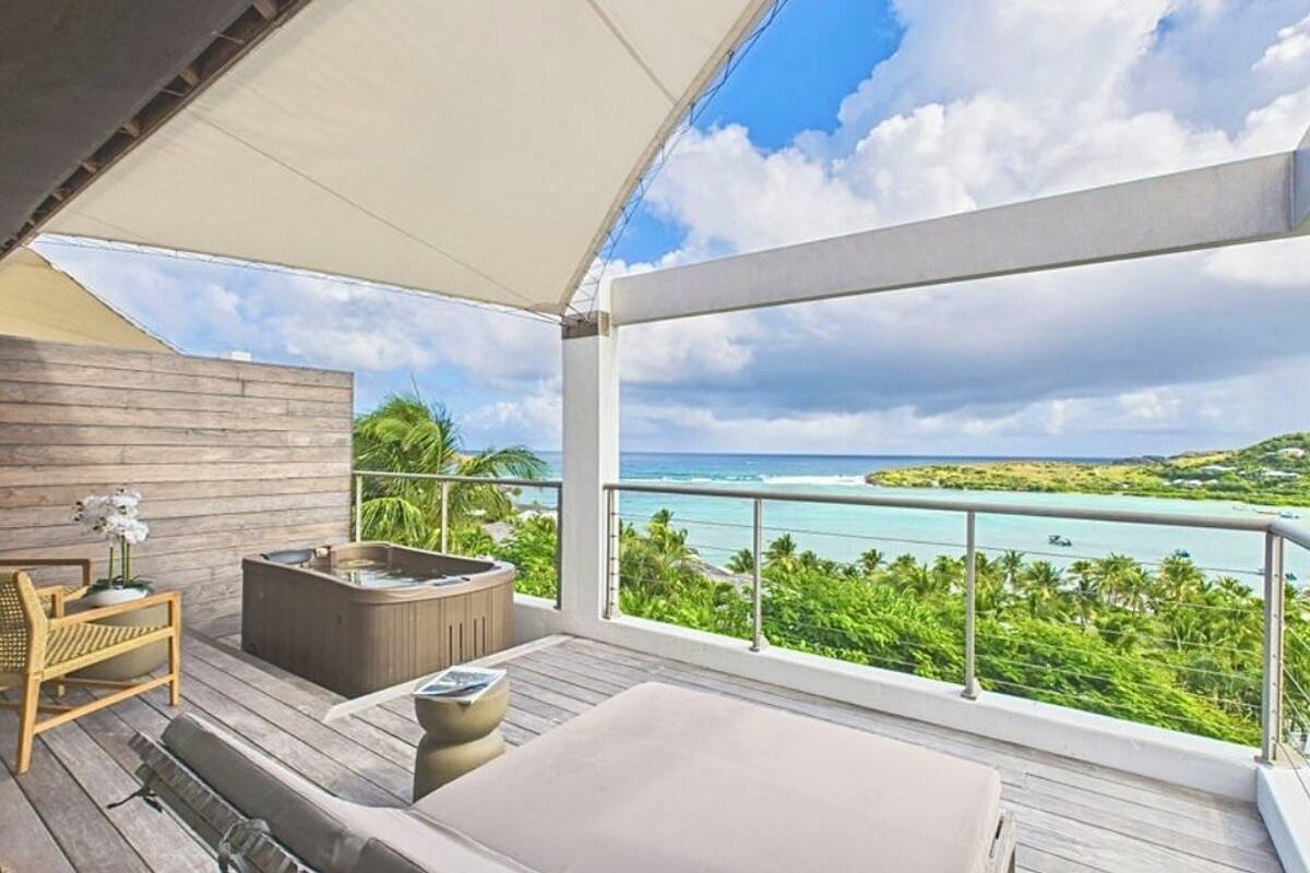 A balcony with a wooden floor, a sofa, a chair, a hot tub, and a view of a turquoise sea and green landscape.